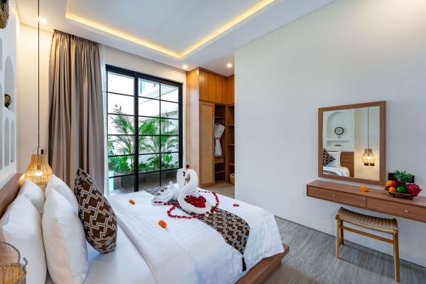 Romantic Bali-style hotel bedroom with towel swan and rose petals on king bed, tropical garden view through sheer-curtained windows, wooden vanity with fruit bowl.